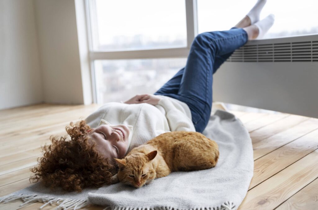 Dans cette scène de pure détente, une femme se repose confortablement sur le sol, son chat blotti près d'elle. Elles profitent toutes deux de la douce chaleur émanant du chauffage au sol, créant un moment de quiétude et de complicité. Le sol devient un oasis de bien-être, où le temps s'étire lentement, et où la chaleur enveloppe leurs corps et leurs âmes. Les sourires apaisés sur leurs visages témoignent de l'amour et de la quiétude partagés, créant une harmonie parfaite entre l'humain et son fidèle compagnon à quatre pattes.