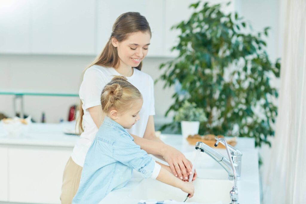 Dans cette scène chaleureuse, une mère attentionnée et sa petite fille partagent un moment de complicité. Elles se tiennent devant un évier rempli d'eau chaude, où la vapeur monte doucement, créant une ambiance apaisante. La mère guide doucement la main de sa fille, lui montrant comment utiliser l'eau chaude en toute sécurité. C'est un instant précieux de transmission de connaissances et de tendresse entre générations, un moment où l'amour et le savoir-faire se partagent, créant des souvenirs inoubliables.